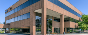 Exterior of the Work Simple Westminster coworking location, a multi-story brick office building with floor-to-ceiling windows and a covered entrance, surrounded by mature trees under a clear blue sky.