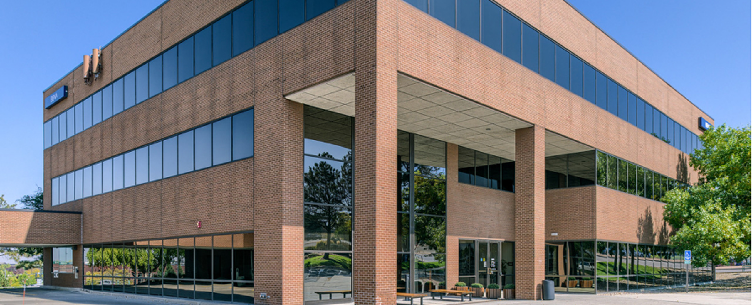 Exterior of the Work Simple Westminster coworking location, a multi-story brick office building with floor-to-ceiling windows and a covered entrance, surrounded by mature trees under a clear blue sky.