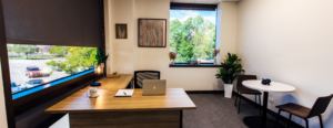 A furnished private office at Work Simple with a wood-topped desk, laptop, and Work Simple coffee mug, a small round meeting table with two chairs, and large windows overlooking a tree-lined parking area.