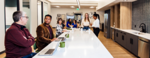 Professionals gathered around a long white kitchen island at a Work Simple coworking location, with laptops and coffee mugs on the counter and glass-walled offices visible in the background.