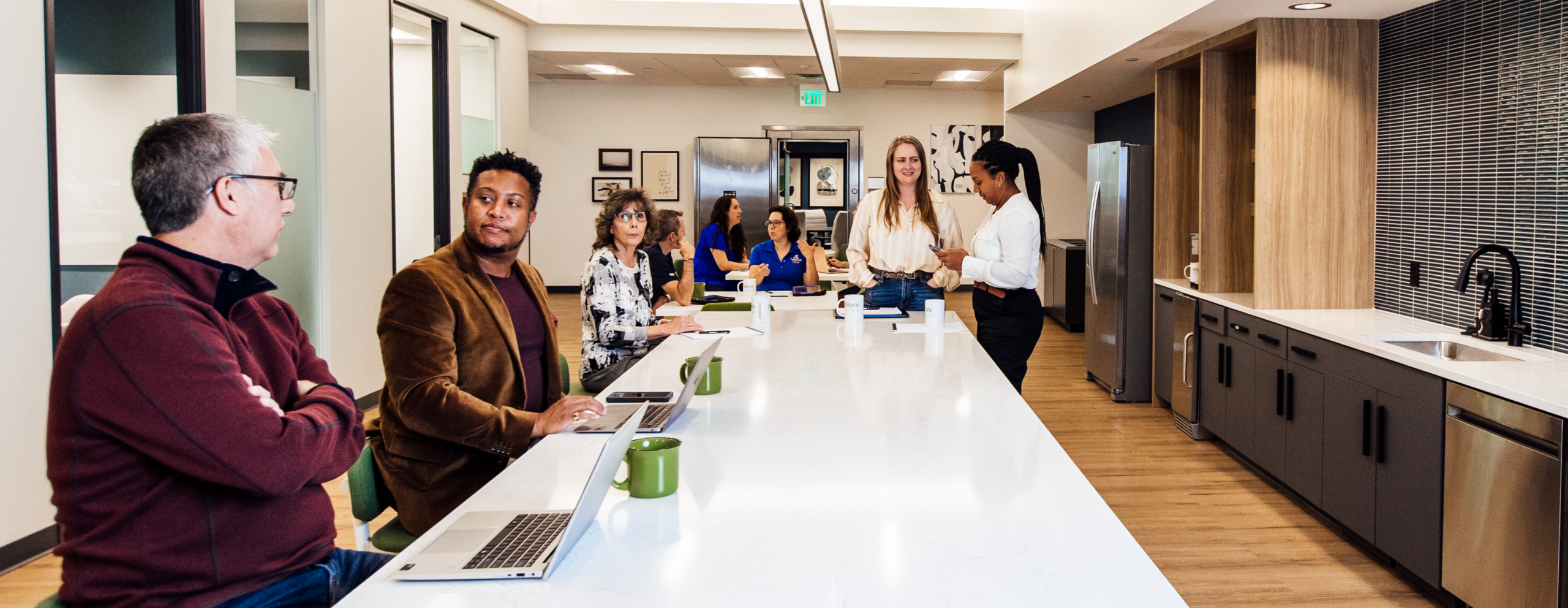 Professionals gathered around a long white kitchen island at a Work Simple coworking location, with laptops and coffee mugs on the counter and glass-walled offices visible in the background.