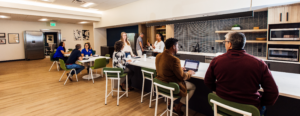 Wide view of the Work Simple common area and kitchen, with members working at bar-height seating along a white island, a small group meeting at a nearby table, and modern finishes including wood cabinetry and tile backsplash throughout.