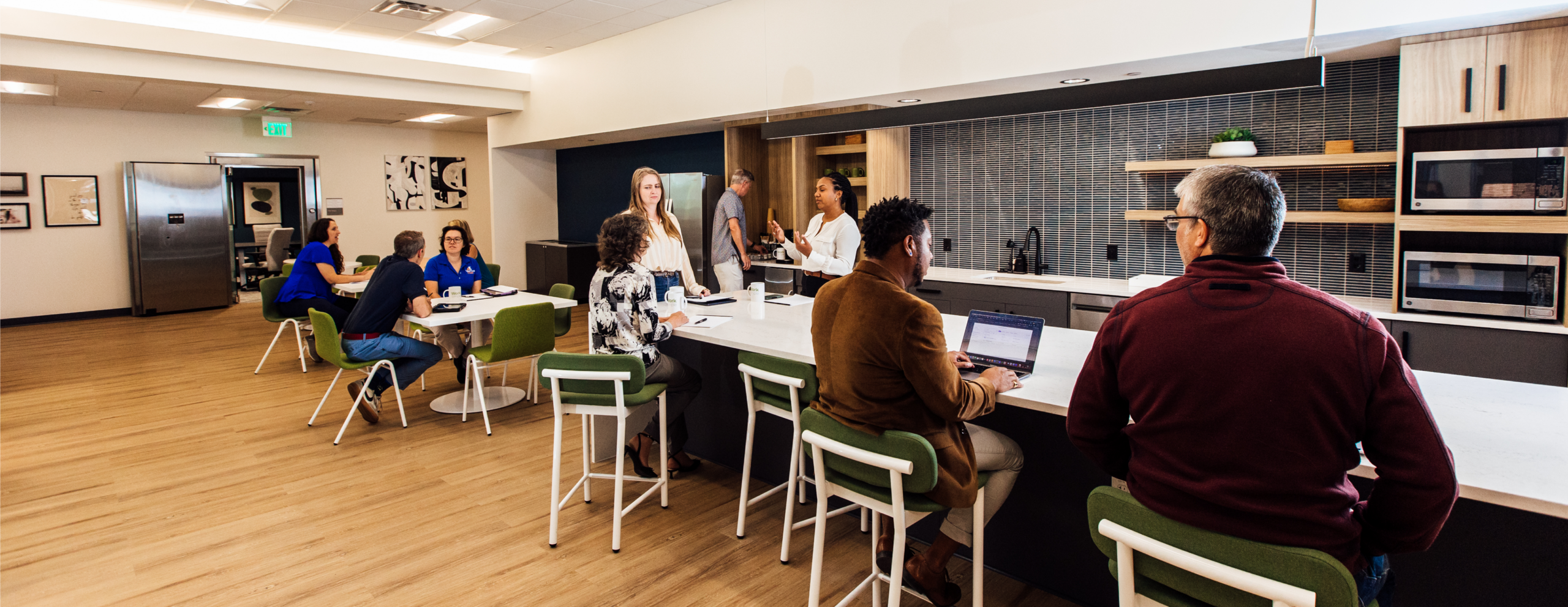 Wide view of the Work Simple common area and kitchen, with members working at bar-height seating along a white island, a small group meeting at a nearby table, and modern finishes including wood cabinetry and tile backsplash throughout.
