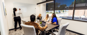Business professionals sitting and talking around a meeting room table while someone writes on a white board