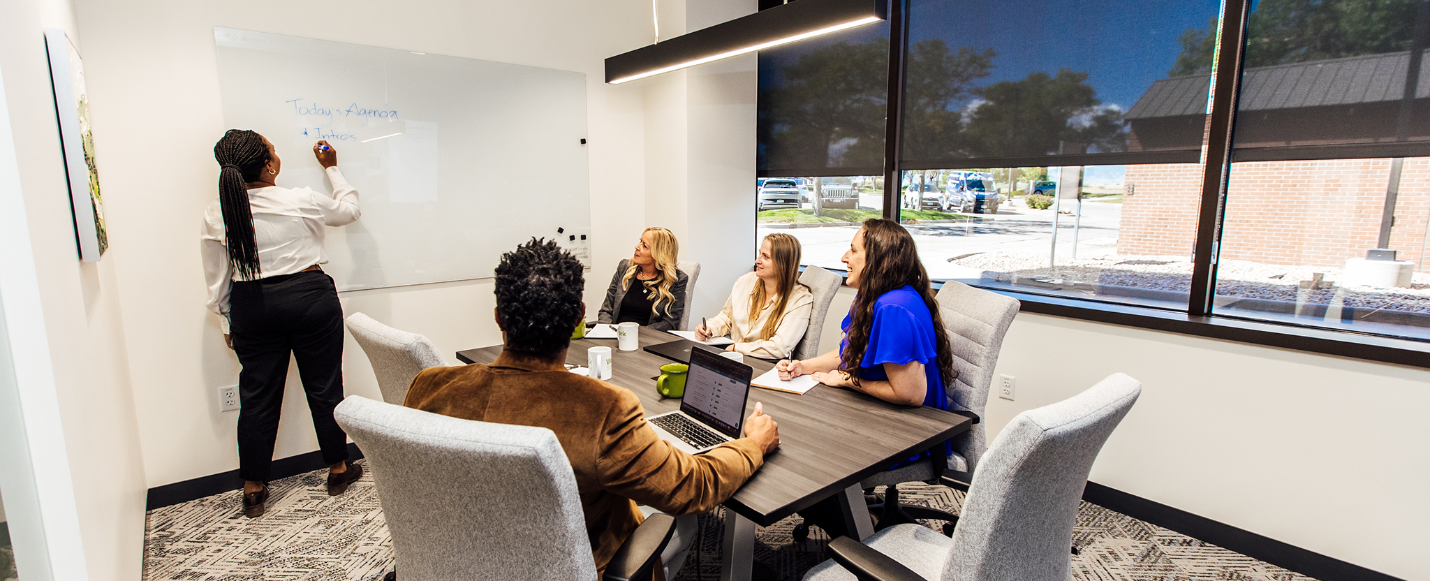 Business professionals sitting and talking around a meeting room table while someone writes on a white board