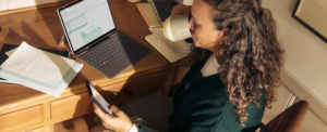 A woman sitting at a desk at home while drinking coffee and looking at her phone