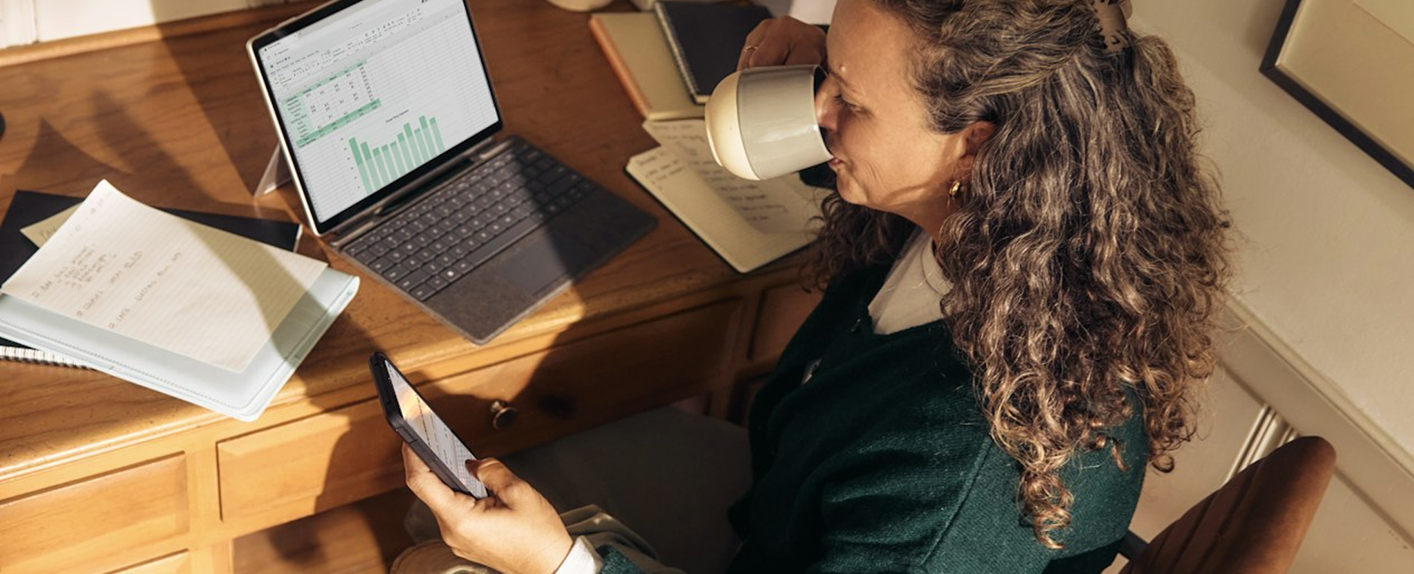 A woman sitting at a desk at home while drinking coffee and looking at her phone
